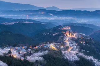 Aerial view of a mountain village at dusk, with illuminated streets winding among cherry blossom trees, surrounded by forested hills and mist, and distant mountains in the background.