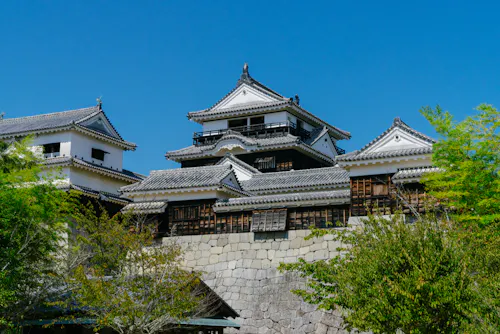 A traditional Japanese castle with white walls, dark tiled roofs, and wooden accents stands behind a stone wall, surrounded by green trees under a clear blue sky.