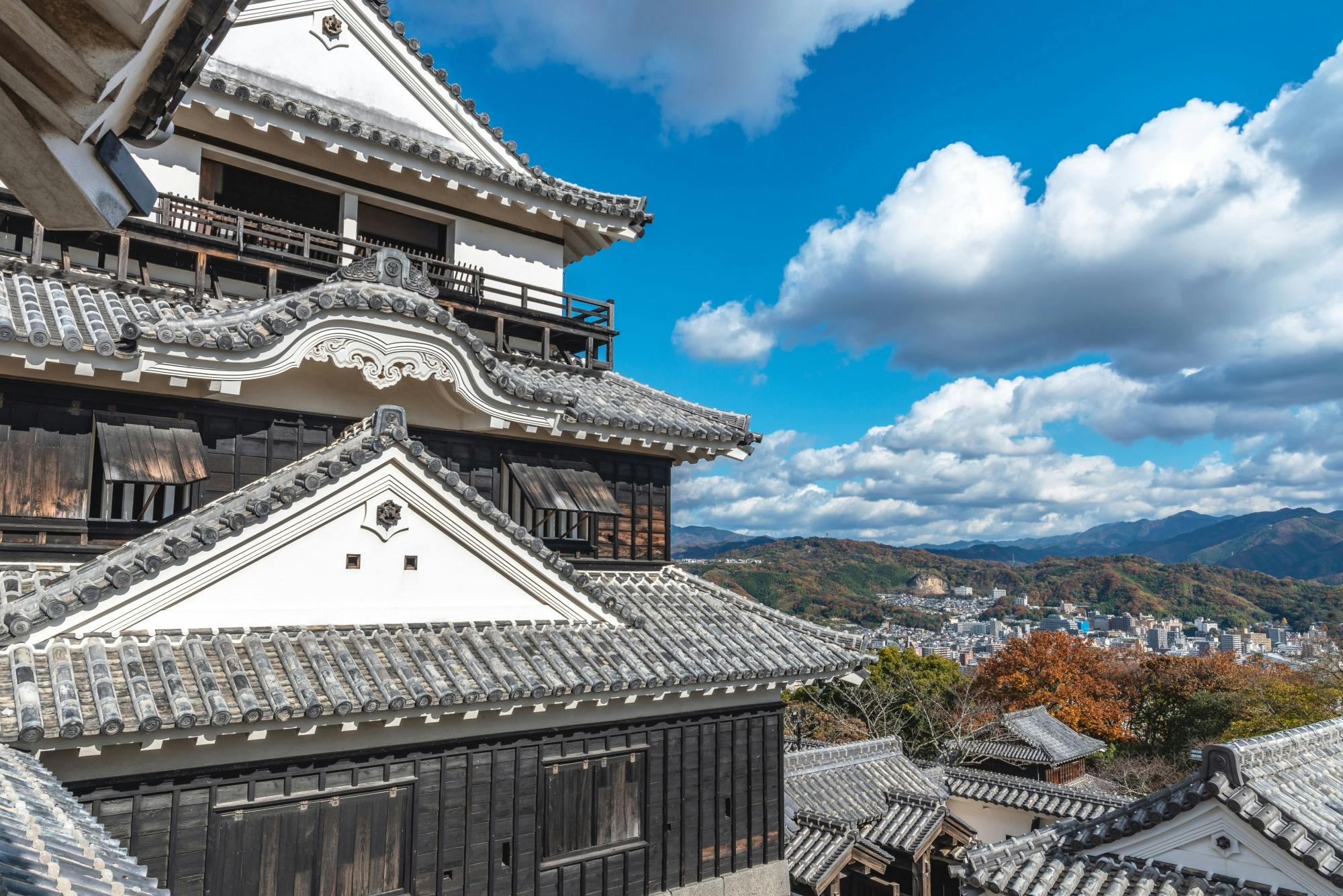 Traditional Japanese castle with white walls and grey tiled roofs, set against a backdrop of a city, colorful autumn trees, rolling hills, and a blue sky with clouds.