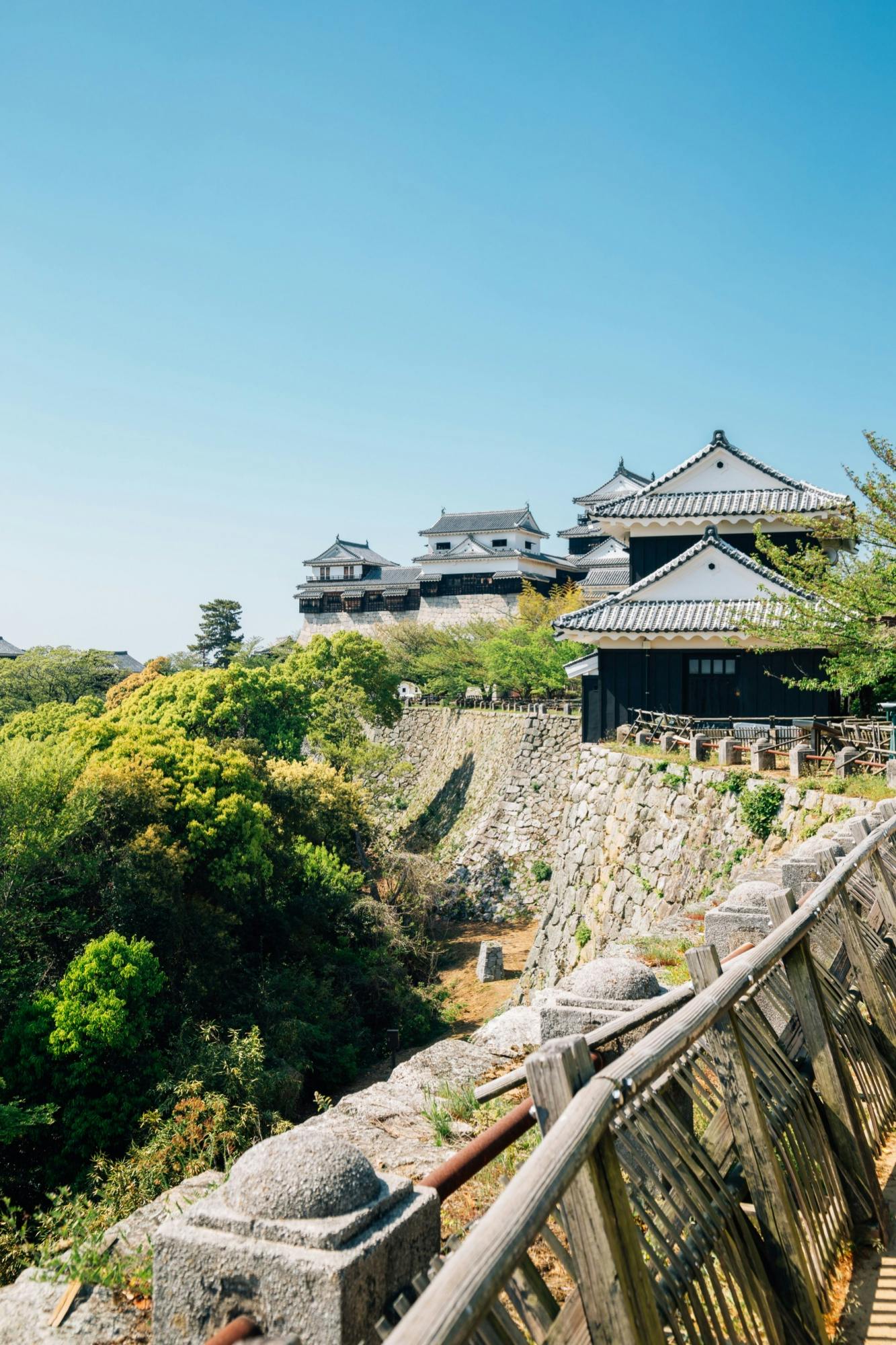 A traditional Japanese castle with black rooftops sits atop a stone wall surrounded by lush green trees under a clear blue sky. A wooden fence runs along the foreground.