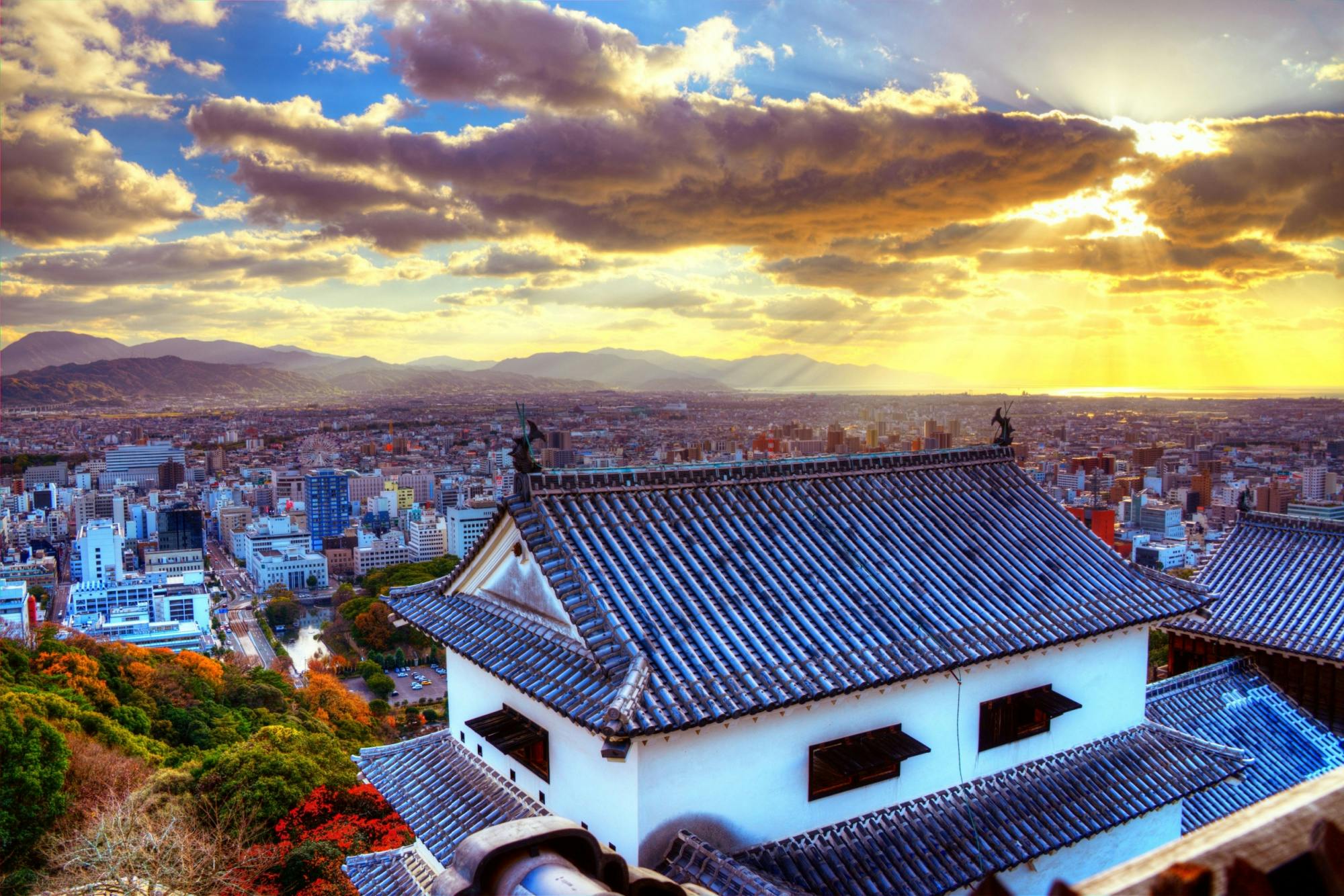 A scenic cityscape view from a traditional Japanese castle roof with blue tiles, overlooking a city at sunset. Sun rays stream through dramatic clouds, illuminating the mountains and colorful autumn trees below.