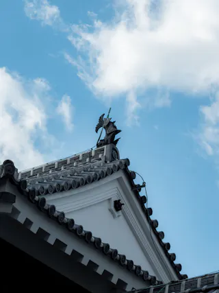A traditional Japanese roof with decorative tiles and a black fish-shaped ornament at the corner, set against a blue sky with scattered white clouds.