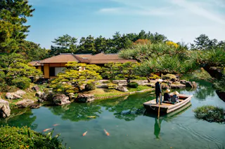 A small wooden boat with three people floats on a tranquil pond surrounded by manicured trees and a traditional Japanese building, with colorful koi fish visible in the clear green water.