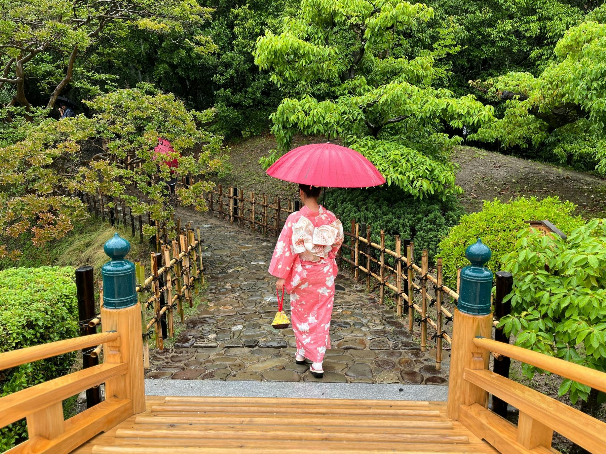 A person in a pink floral kimono holds a matching pink umbrella while walking down a stone path in a lush, green Japanese garden. Wooden railings border the path, and trees surround the scene.