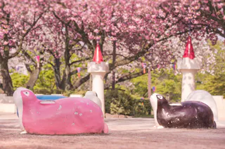 Two large bunny-shaped playground rides, one pink and one black, sit under blooming cherry blossom trees. White castle-like structures with red cone roofs are visible in the background.