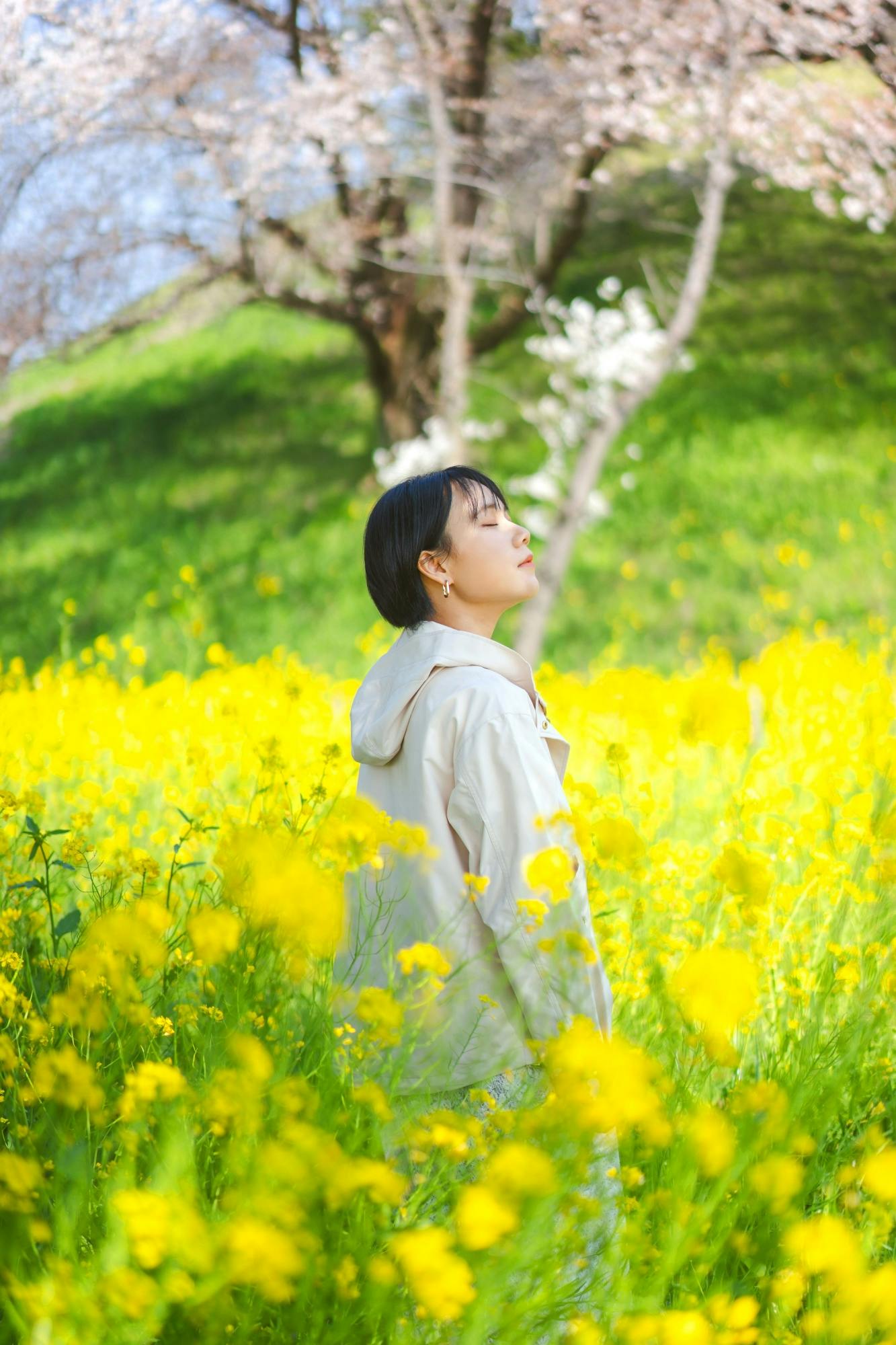 A young woman stands in a field of vibrant yellow flowers, eyes closed, enjoying the sunlight. Green grass and blooming trees with white blossoms are in the background.