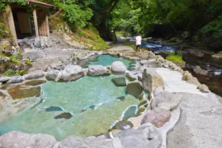 A natural hot spring pool with clear blue water surrounded by rocks and lush greenery. A person in a white shirt walks on a stone path beside a river, with a wooden structure visible on the left.