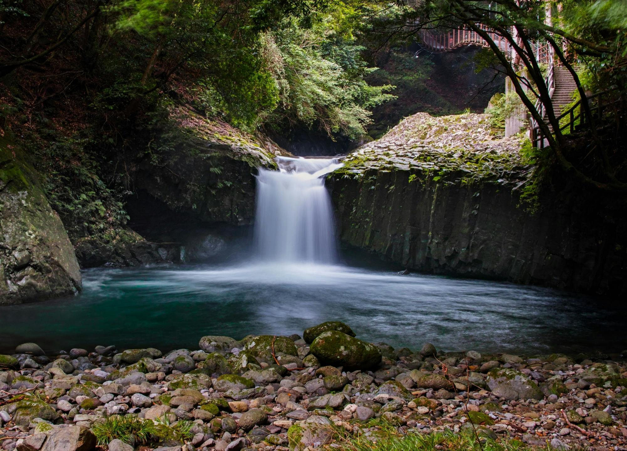 A small waterfall flows into a serene, clear pool surrounded by mossy rocks and lush green trees, with a rocky shoreline in the foreground and a wooden walkway visible in the upper right corner.