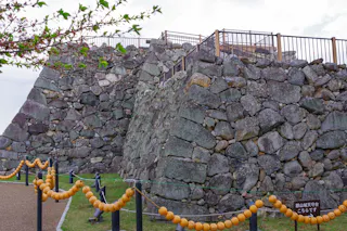 A large stone wall with rough, stacked rocks forms part of a historic castle structure. Wooden railings line the top, and orange rope barriers with balls enclose the area. A small sign sits near the base.