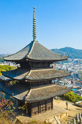 A traditional Japanese three-story pagoda with tiled roofs stands on a hillside, overlooking a city and distant green mountains under a clear blue sky.