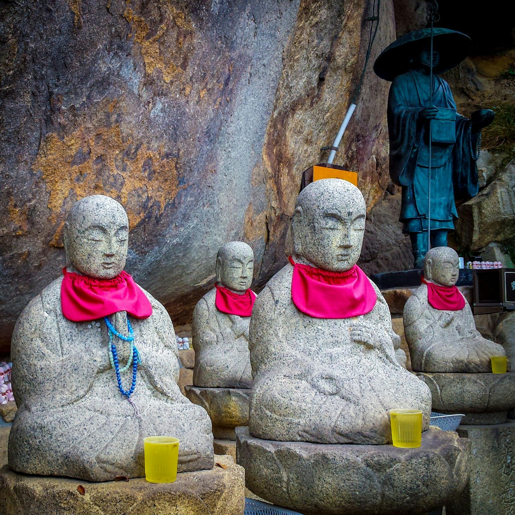 Several stone Buddha statues wearing bright pink bibs are seated in a row with yellow cups placed in front of them. A larger dark statue stands in the background against a rocky wall.