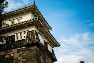 A traditional Japanese castle tower with white walls, wooden accents, and a tiled roof, set against a blue sky with scattered clouds.