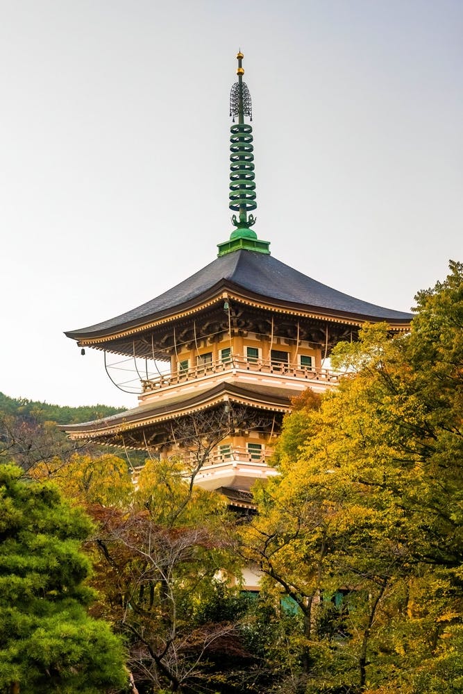 A traditional Japanese pagoda with a dark roof and green spire rises above colorful autumn trees, set against a clear sky and distant wooded hills.