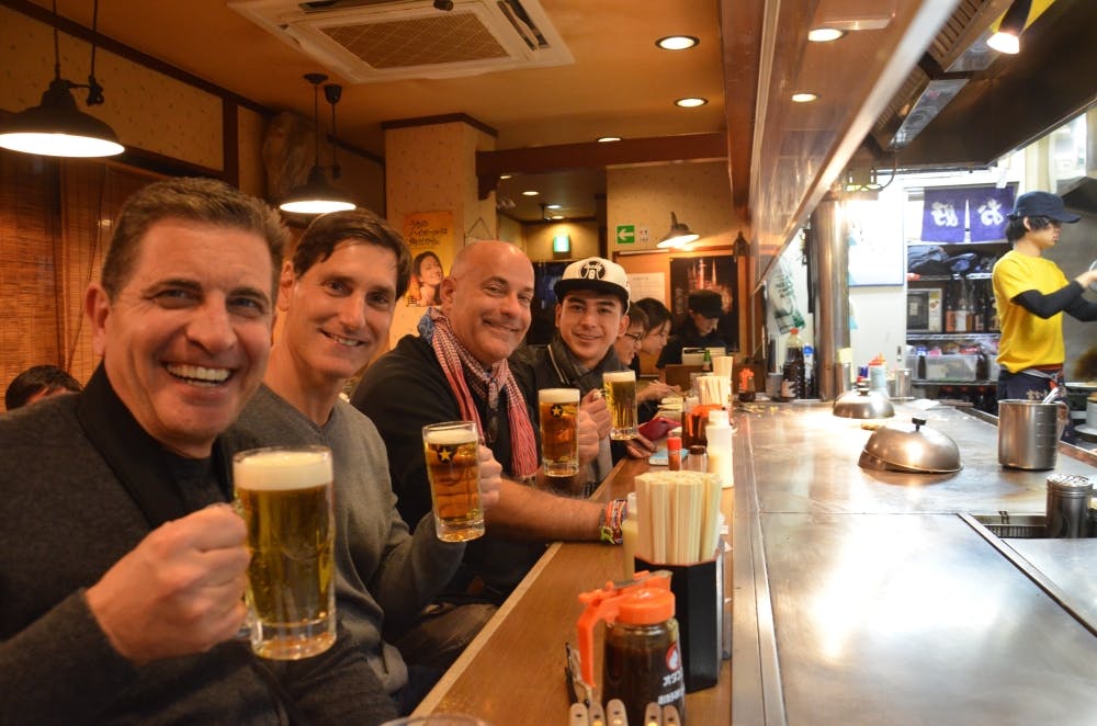 Four smiling men sit at a bar counter, each holding a glass of beer. The setting appears to be a cozy, casual restaurant with wooden decor and other patrons in the background.
