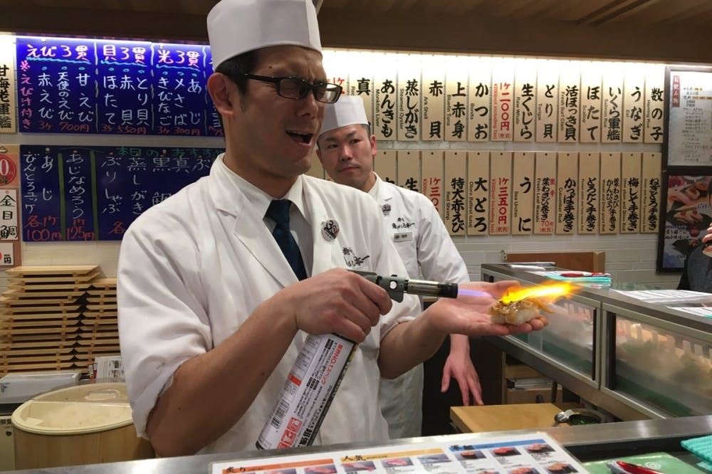 A sushi chef using a blowtorch to sear sushi in his hand, while another chef stands in the background. Both are wearing white uniforms and hats, with menus in Japanese on the wall behind them.