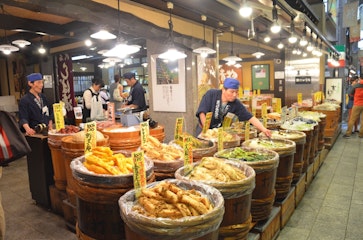 Shopkeepers in blue uniforms arrange large wooden barrels filled with various pickled vegetables at an indoor Japanese market. Bright yellow signs with prices are displayed on each barrel under hanging lights.