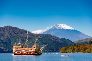 A colorful pirate-style ship sails on a lake surrounded by wooded hills, with Mount Fuji, its summit capped with snow, rising majestically in the background under a clear blue sky.