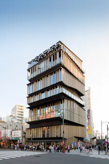 A modern, multi-story building with glass and vertical wooden slats stands on a busy street corner, surrounded by people and urban structures under a clear blue sky.