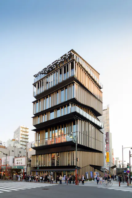 A modern, multi-story building with glass and vertical wooden slats stands on a busy street corner, surrounded by people and urban structures under a clear blue sky.