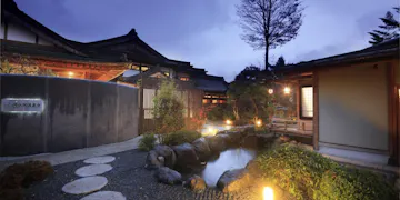 Fuji Onsenji Yumedono Traditional Japanese inn at dusk with wooden buildings, paper lanterns, a small pond surrounded by rocks and plants, and a stone path leading through a peaceful garden. Soft lights illuminate the serene scene.