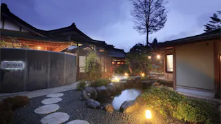 Traditional Japanese inn at dusk with wooden buildings, paper lanterns, a small pond surrounded by rocks and plants, and a stone path leading through a peaceful garden. Soft lights illuminate the serene scene.