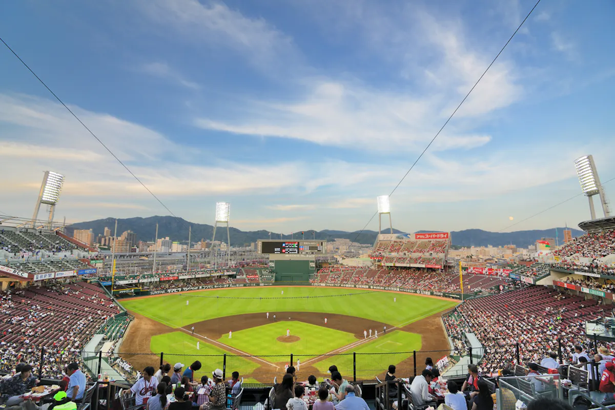 An NPB crowd at dusk, where organized cheering runs the length of the game A packed open-air Japanese baseball stadium at dusk with fans in the stands and mountains behind the outfield