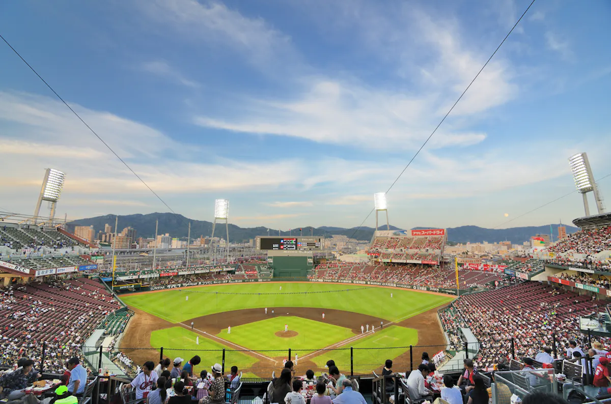 A packed open-air Japanese baseball stadium at dusk with fans in the stands and mountains behind the outfield