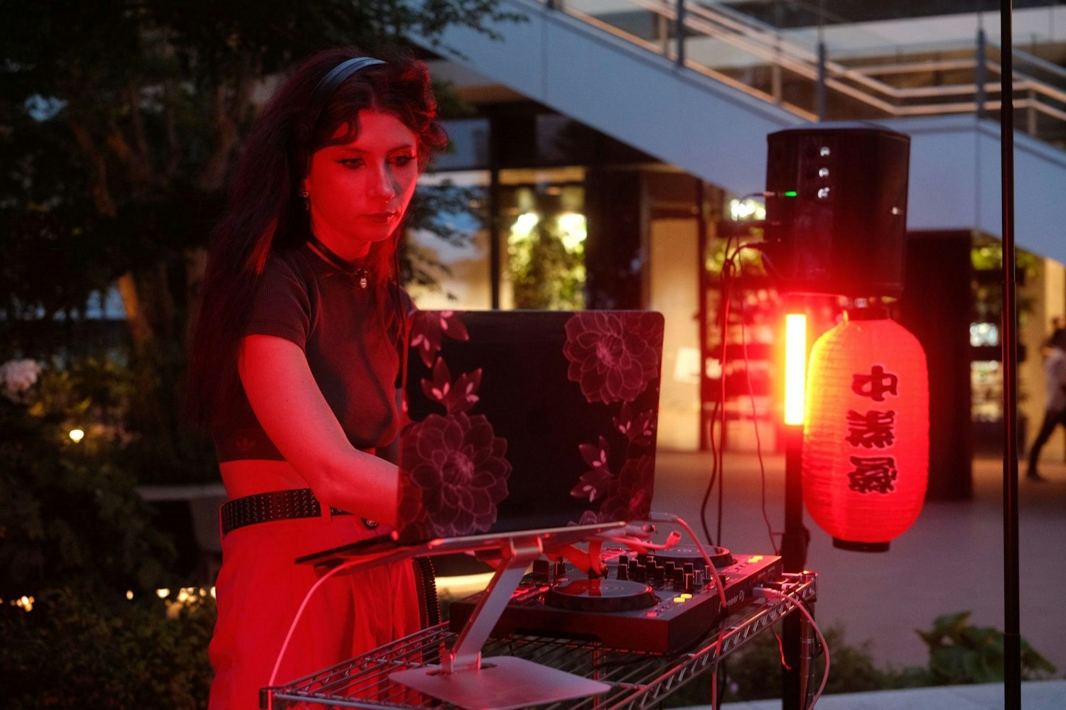 A woman DJ, lit by red light, mixes music on a laptop and DJ controller. A glowing lantern with Chinese characters hangs nearby. She is outdoors, surrounded by modern architecture and greenery.