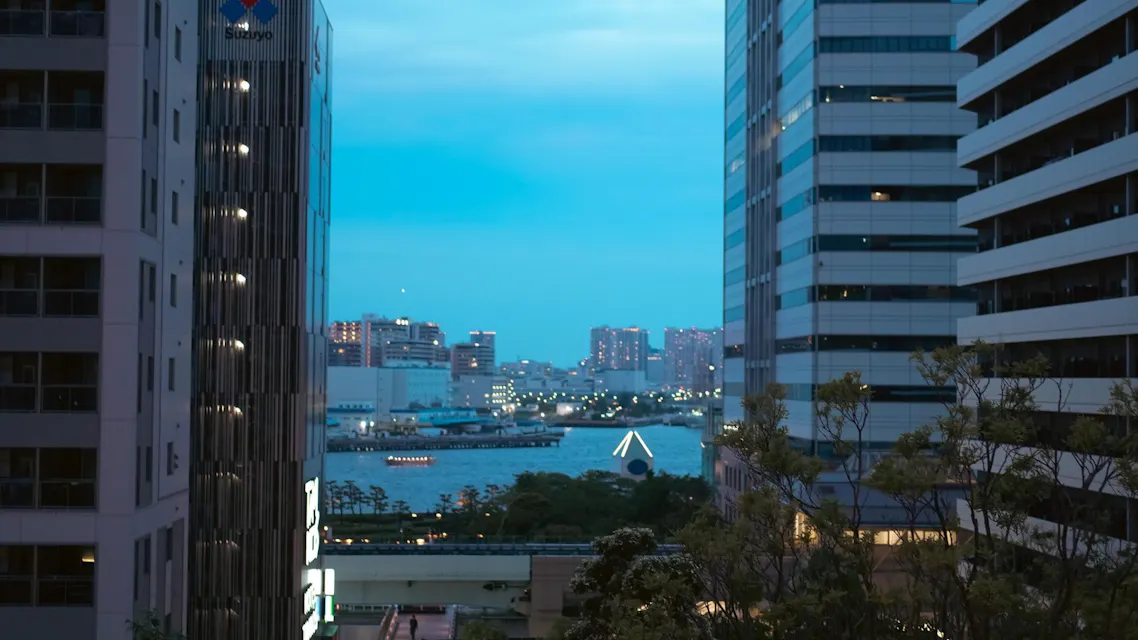 A cityscape at dusk with tall buildings framing a river. Lights from the windows and boats reflect on the water, and more buildings are visible in the background under a blue evening sky.