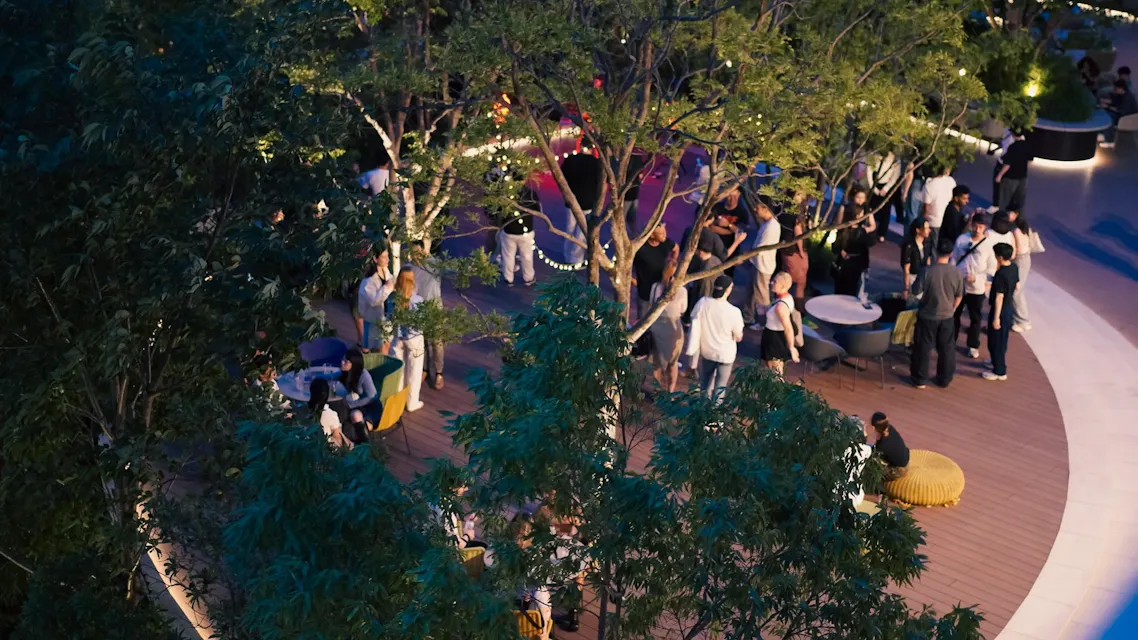 Aerial view of people socializing in an outdoor terrace surrounded by trees and string lights, with groups seated at tables and others standing and chatting in a relaxed, lively atmosphere.