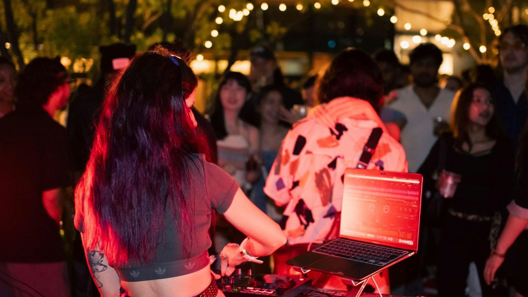 A DJ with long dark hair plays music on a laptop and mixer at an outdoor night event, with a crowd of people dancing and string lights glowing in the background.