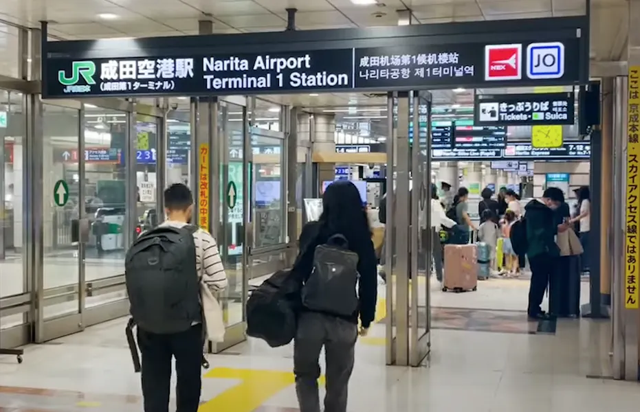 Travelers with backpacks walk towards the entrance of Narita Airport Terminal 1 Station. Signs are displayed in multiple languages, including Japanese and English, and people are visible in the background near ticket machines and gates.
