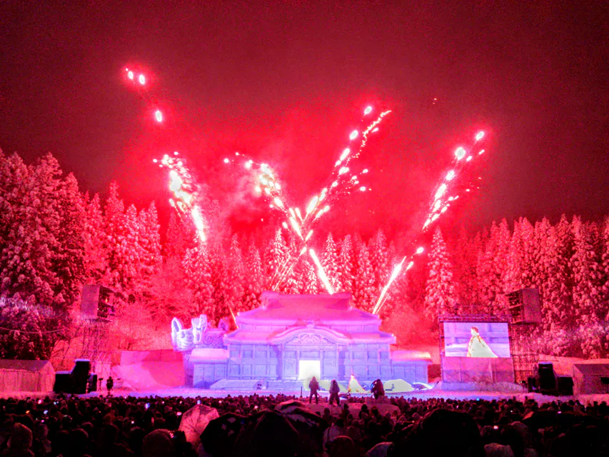 A vibrant display of fireworks illuminates the night sky over an ice sculpture resembling a traditional building at a winter festival. The scene is surrounded by snow-covered trees, with people gathered to watch the spectacle.