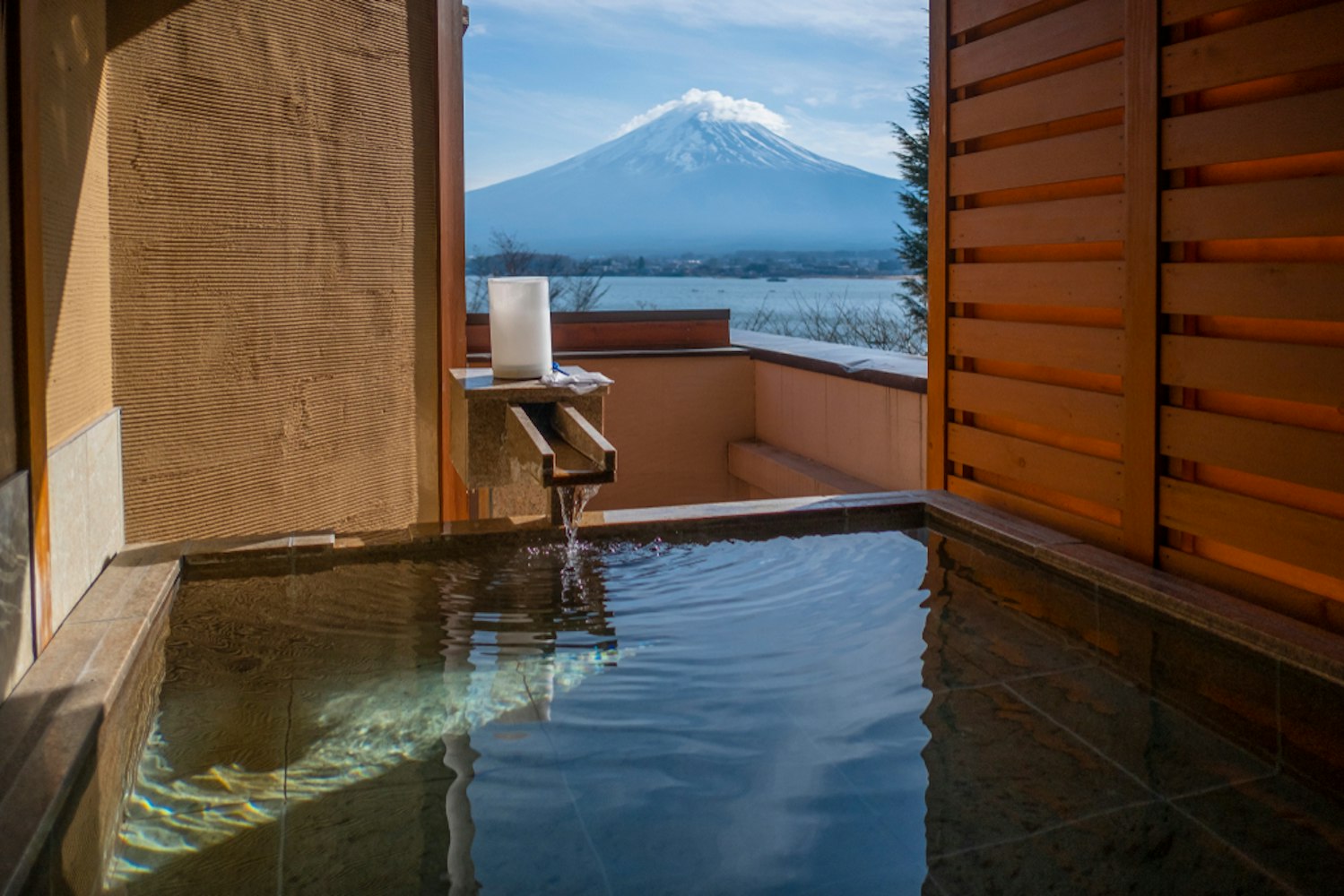 A serene outdoor hot spring bath with flowing water, enclosed by wooden panels, offering a stunning view of a snow-capped mountain and a lake under a clear blue sky.