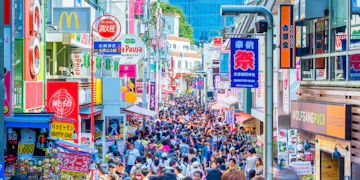 Harajuku A large crowd of people fills a busy shopping street lined with colorful shops, signs, and advertisements in Tokyo, Japan, under bright daylight.