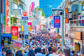 A large crowd of people fills a busy shopping street lined with colorful shops, signs, and advertisements in Tokyo, Japan, under bright daylight.