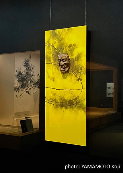 A traditional Japanese mask is mounted on a vibrant yellow panel with abstract patterns, displayed upright in a dimly lit museum exhibit. Photo credit: YAMAMOTO Koji.