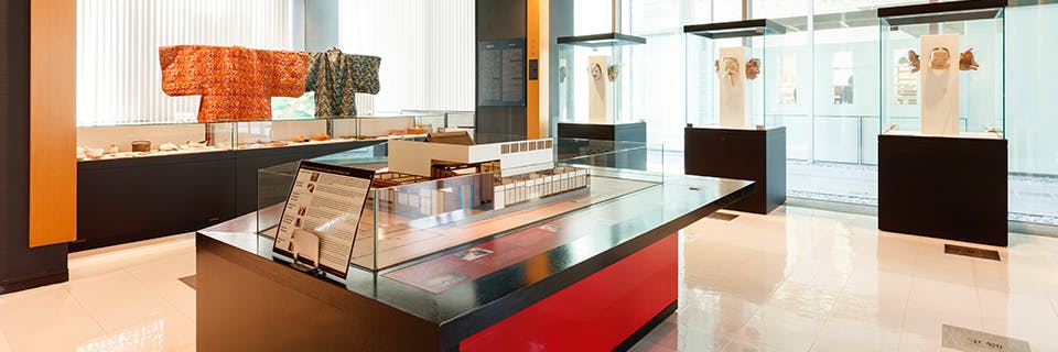 A brightly lit museum exhibit featuring traditional clothing, display cases with artifacts, and a detailed architectural model on a central table, surrounded by large windows and modern decor.
