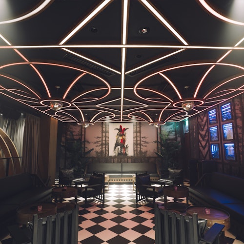 1 Oak Tokyo A stylish lounge with black sofas, round tables, checkered tile floor, and a ceiling featuring geometric light designs. A decorative art piece is displayed at the far end of the room.