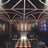 A stylish lounge with black sofas, round tables, checkered tile floor, and a ceiling featuring geometric light designs. A decorative art piece is displayed at the far end of the room.