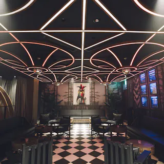A stylish lounge with black sofas, round tables, checkered tile floor, and a ceiling featuring geometric light designs. A decorative art piece is displayed at the far end of the room.
