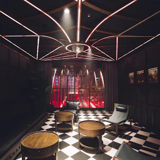 A modern lounge with black-and-white checkered floors, dark walls, circular wooden tables, black chairs, and a ceiling with geometric neon lights. Red lighting highlights an arched window at the far end.