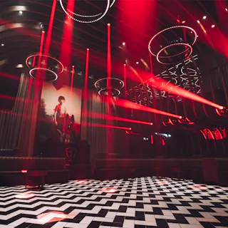 A nightclub interior with black-and-white checkered floor, red lighting, circular ceiling lights, and a large wall art of a person. Booth seating lines the walls, and the space is empty of people.