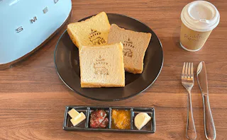 Three slices of thick toast with bakery stamps on a plate, accompanied by small portions of butter, jam, and spreads, with a cup of coffee, fork, and knife on a wooden table near a blue toaster.