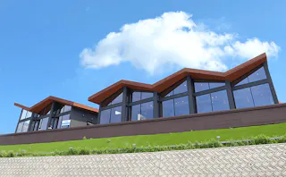 Modern building with large glass windows and a zigzag roofline, set on a grassy slope with a patterned retaining wall, under a bright blue sky with a few clouds.