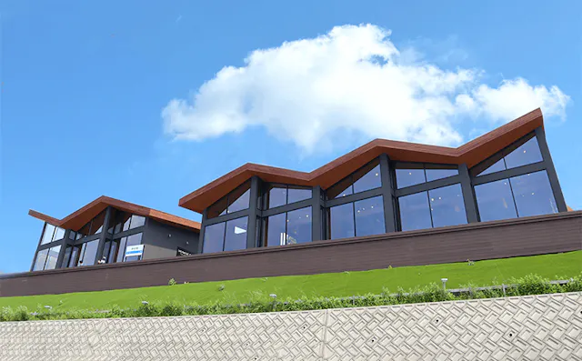 Modern building with large glass windows and a zigzag roofline, set on a grassy slope with a patterned retaining wall, under a bright blue sky with a few clouds.