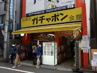 A yellow storefront in Japan with Japanese signage above the entrance, displaying capsule toy vending machines inside. Several people are entering and browsing, while others walk past on the street.