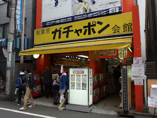 A yellow storefront in Japan with Japanese signage above the entrance, displaying capsule toy vending machines inside. Several people are entering and browsing, while others walk past on the street.