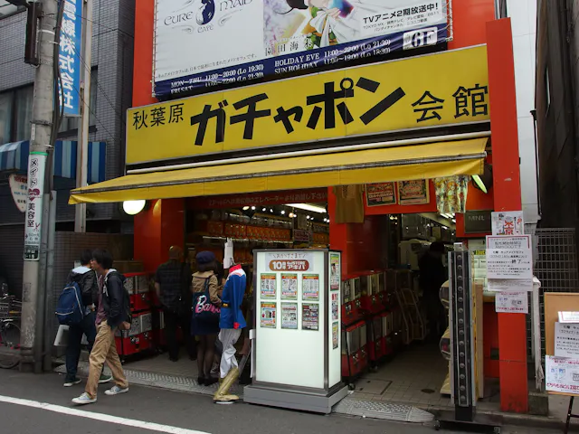 A yellow storefront in Japan with Japanese signage above the entrance, displaying capsule toy vending machines inside. Several people are entering and browsing, while others walk past on the street.