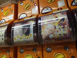 Close-up of orange Japanese capsule toy vending machines (gachapon) with display cards showing face-themed toy advertisements and price labels for 300 yen.